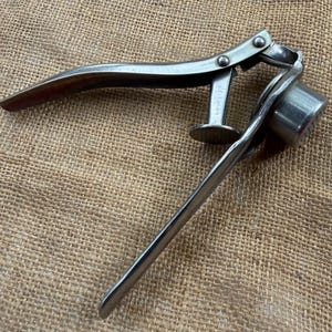 May include: A silver-colored metal nutcracker with two long, curved handles connected by a hinge. The tool features a cylindrical cup for holding a nut and a flat, circular plunger mechanism that presses down when the handles are squeezed together.