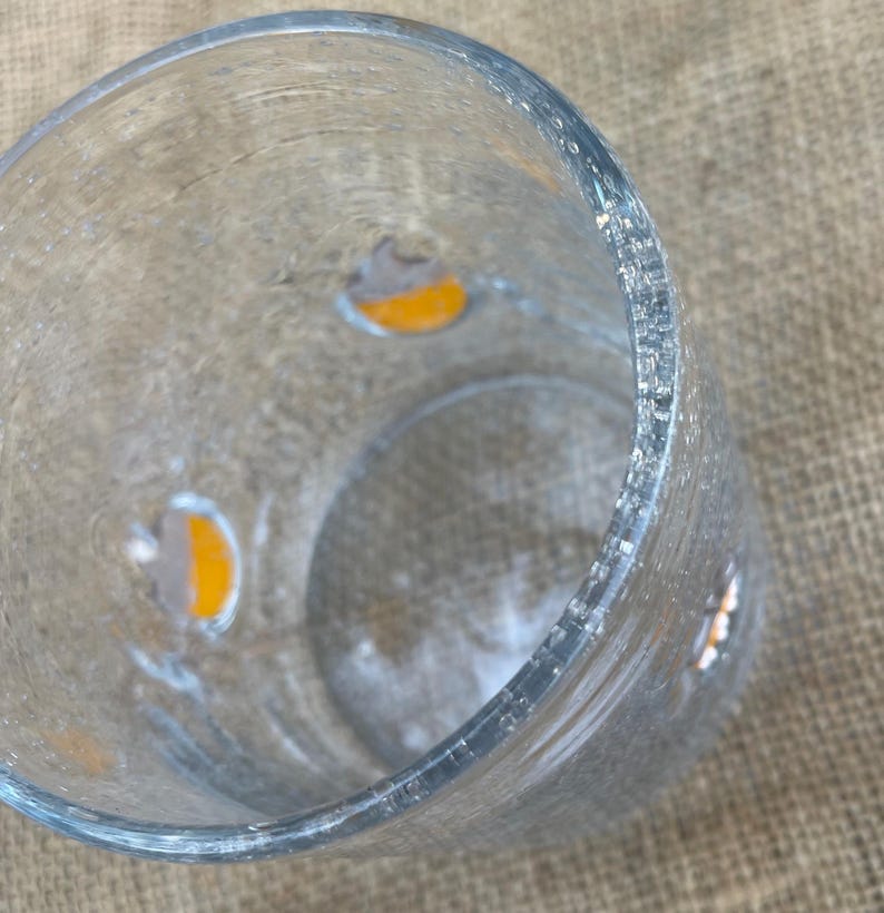 May include: A clear, cylindrical glass vase with a textured surface. The vase has small, irregular orange and gray accents. The rim of the vase is slightly uneven. The vase is empty.