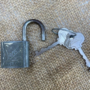 Puede incluir: Un candado de metal vintage abierto, con la palabra "Slaymaker" y "RUSTLESS" en relieve en la parte delantera. Dos llaves con la palabra "STALOCK" están junto al candado. El candado y las llaves son plateados y están sobre una superficie beige texturizada.
