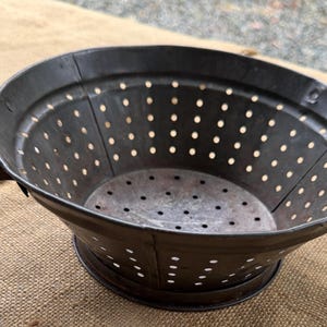 May include: A dark brown metal colander featuring a wide, circular rim, two curved side handles, and a series of evenly spaced drainage holes throughout the bowl and base. The surface shows a weathered, matte finish with visible signs of age.