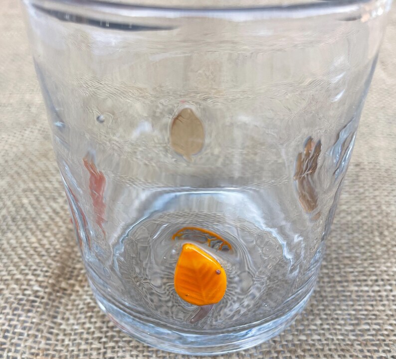 May include: A clear glass tumbler with a textured surface. The glass features decorative leaf-shaped accents in shades of orange and brown. The tumbler is designed for everyday use.