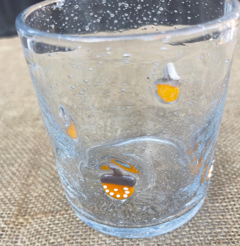 May include: A clear glass tumbler with a textured surface and small bubbles. The glass is decorated with orange, white, and brown acorn designs. The tumbler is suitable for serving beverages.