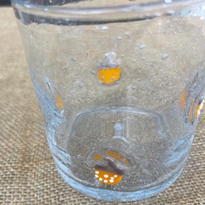 May include: Clear glass tumbler with acorn designs in orange, brown, and white. The glass has a bubbly texture and is partially filled. The acorns are evenly spaced around the glass, adding a decorative touch to the drinkware.