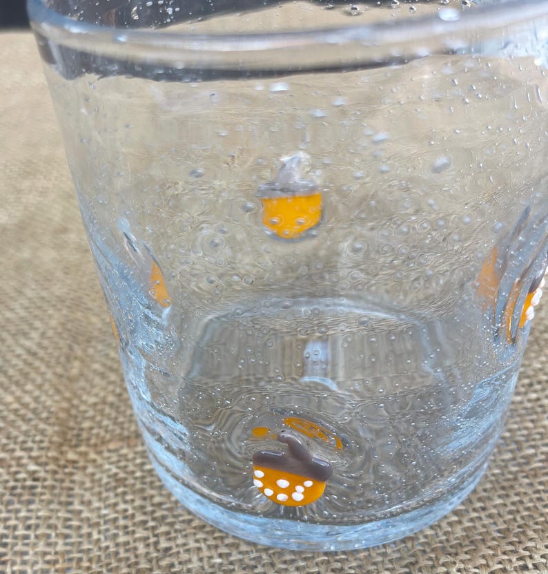 May include: Clear glass tumbler with acorn designs in orange, brown, and white. The glass has a bubbly texture and is partially filled. The acorns are evenly spaced around the glass, adding a decorative touch to the drinkware.