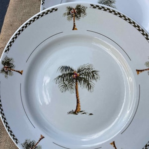 May include: Two white ceramic plates with a tropical palm tree design. Each plate features a central palm tree image and smaller palm tree accents around the rim. The plates have a decorative border with a repeating geometric pattern.