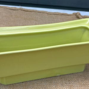 Puede incluir: Una jardinera rectangular de cerámica verde claro con un borde acampanado y una base escalonada. La jardinera tiene un acabado liso y brillante y un diseño moderno. La jardinera mide aproximadamente 30 cm de largo.