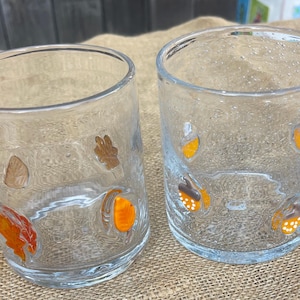 May include: Two clear glass tumblers with autumn-themed decorations. One tumbler features leaf and acorn designs in orange, brown, and tan. The other tumbler has acorn designs in orange, brown, and white. The glasses are on a burlap surface.