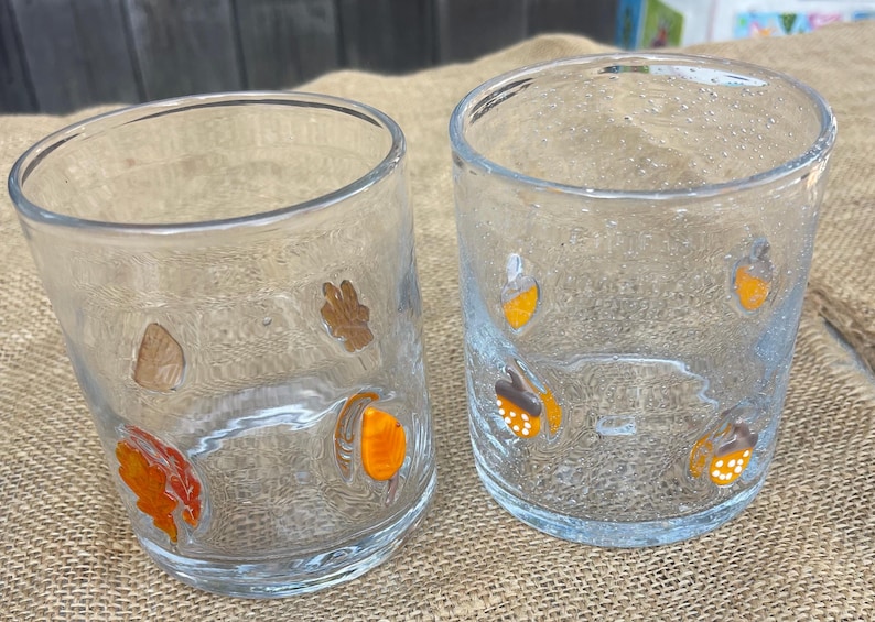May include: Two clear glass tumblers with autumn-themed decorations. One tumbler features leaf and acorn designs in orange, brown, and tan. The other tumbler has acorn designs in orange, brown, and white. The glasses are on a burlap surface.
