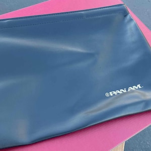 Op de afbeelding: Een zwarte, rechthoekige etui met het zilveren "PAN AM" logo. De etui ligt op een roze ondergrond. De etui lijkt gemaakt van een glad, mogelijk synthetisch materiaal.