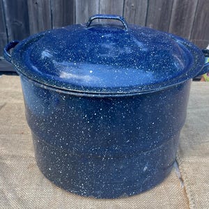 May include: A large, blue enamelware pot with a lid. The pot is speckled with white spots and has two side handles and a handle on the lid. The pot is sitting on a burlap surface.