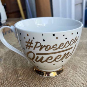 May include: White ceramic teacup with a gold handle and base. The cup features the text "#Prosecco Queen" in gold lettering, along with gold and black polka dots. A stylish and fun addition to any kitchen.