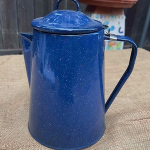 May include: A vintage blue enamelware coffee pot with a lid and handle. The pot has a speckled pattern and a spout for pouring. The handle is attached to the side of the pot. The pot is sitting on a burlap surface.