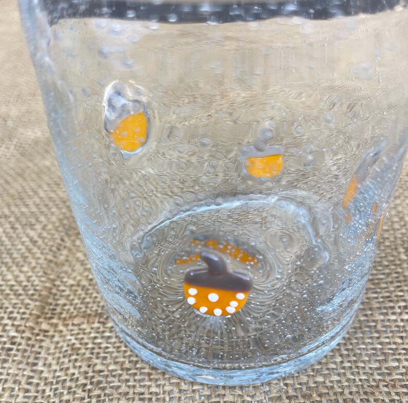 May include: Clear glass tumbler with a textured surface and small bubbles. The glass is decorated with orange and brown acorn designs with white polka dots. The tumbler is likely for beverages.