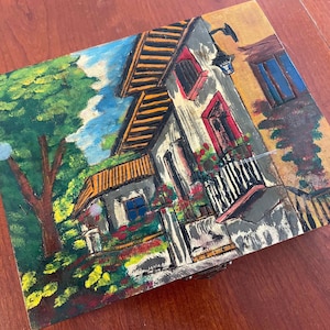 Può includere: Scatola di legno dipinta a mano con una scena colorata di una casa con balcone, circondata da alberi e fiori. La scatola ha un coperchio incernierato e un chiavistello.