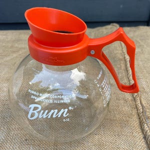 May include: A clear glass Bunn B-12 coffee carafe with an orange plastic lid and handle. The carafe has the Bunn logo and text that reads "BUNN-O-MATIC CORPORATION SPRINGFIELD, ILLINOIS". The carafe is sitting on a burlap surface.