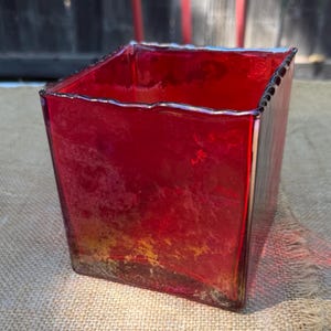 May include: A square glass vase featuring a deep red hue with a mottled, textured finish. The vessel has a straight-sided design with a slightly uneven, organic rim, showcasing a translucent quality that allows light to pass through the glass.