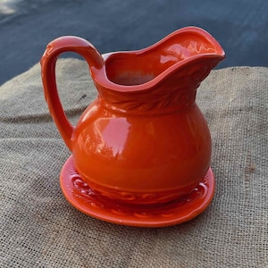 May include: A bright orange ceramic pitcher with a matching saucer. The pitcher has a wide mouth and a curved handle. The saucer has a decorative design around the edge.