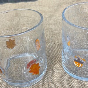 May include: Two clear drinking glasses with textured surfaces. Each glass is decorated with colorful fall-themed images, including acorns and leaves. The glasses are set on a neutral-colored surface.