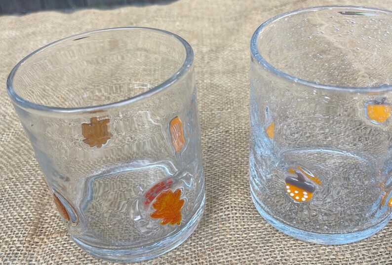 May include: Two clear drinking glasses with textured surfaces. Each glass is decorated with colorful fall-themed images, including acorns and leaves. The glasses are set on a neutral-colored surface.