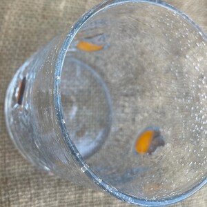 May include: A clear, textured glass with a cylindrical shape. The glass has a slightly flared rim and appears to have small, irregular bubbles on the surface. There are two orange-colored shapes visible inside the glass. The glass is sitting on a tan surface.