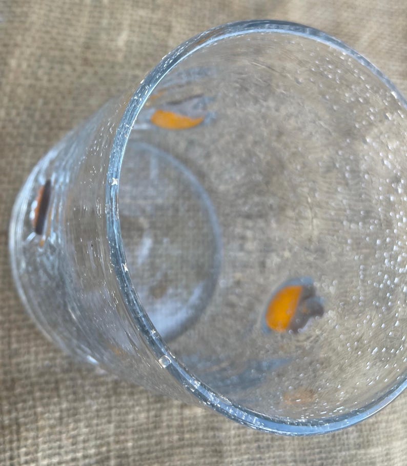 May include: A clear, textured glass with a cylindrical shape. The glass has a slightly flared rim and appears to have small, irregular bubbles on the surface. There are two orange-colored shapes visible inside the glass. The glass is sitting on a tan surface.