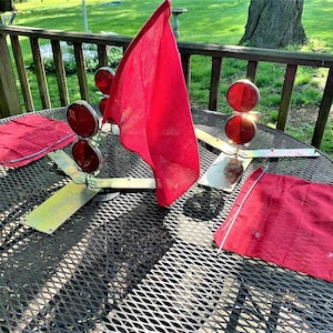 以下が含まれることがあります： 黒い金属製のテーブルに、ヴィンテージの赤い旗と信号灯のコレクションが展示されています。旗は質感のある赤い布でできています。信号灯には2つの赤いレンズがあり、金属製のベースに取り付けられています。