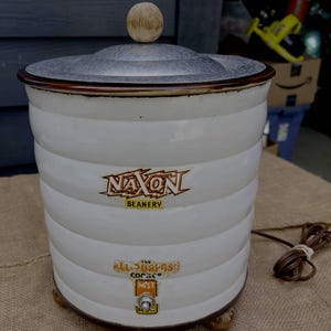 May include: A vintage white and brown Naxon Beanery electric cooker with a lid and wooden knob. The cooker has a ribbed design and features the text "Naxon Beanery" and "The All-Purpose Cooker".
