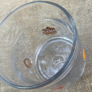 May include: Clear glass drinking glass with a textured surface. The glass has a decorative brown floral design on the bottom and a small orange accent on the side. The glass is empty.