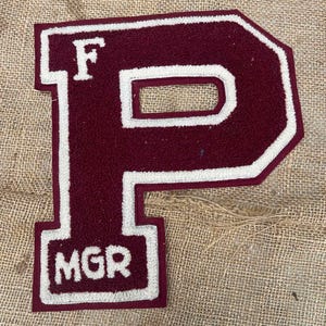 May include: Maroon felt letter "P" with white trim and the letters "F" and "MGR" inside. The letter is a varsity-style patch, likely for a jacket or apparel item. The background is a textured, tan fabric.
