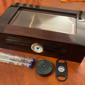 Puede incluir: Un humidor de madera oscura con tapa de vidrio, higrómetro incorporado y revestimiento de cedro. El humidor está abierto y muestra una tapa de vidrio transparente y un revestimiento de cedro. El humidor está sobre una superficie de madera. Hay dos tubos de cristales humidificadores y un cortador de cigarros al lado del humidor.