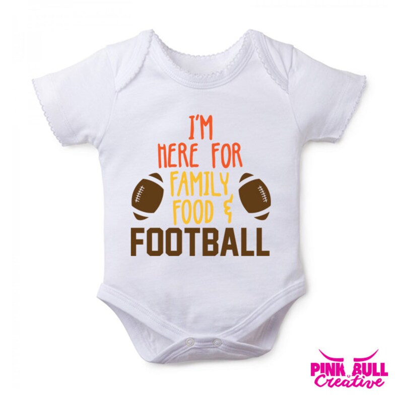 Op de afbeelding: Witte baby romper met bruine voetballen aan weerszijden van de tekst "Ik ben hier voor familie, eten & voetbal".