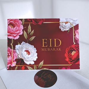 May include: A burgundy Eid Mubarak greeting card with gold detailing and floral illustrations in pink, red, and white. The card is accompanied by a white envelope and a round sticker with the words "With Love" in a script font.