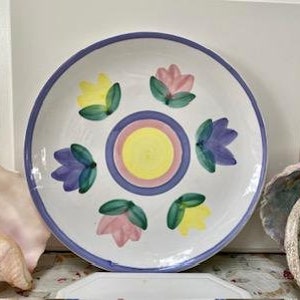 May include: A white ceramic plate with a lavender rim and a central design of concentric circles in yellow, pink, and blue. The plate features six stylized flower designs in yellow, pink, and purple with green leaves.