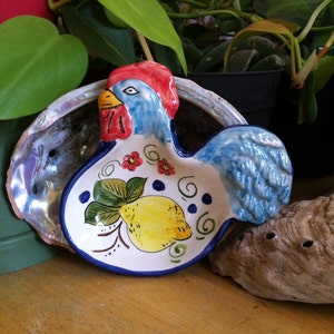 May include: A ceramic rooster-shaped spoon rest. The spoon rest is white with a blue rim and features a yellow lemon and red flower design. The rooster has a red comb, blue feathers, and a yellow beak. Decorative kitchenware.