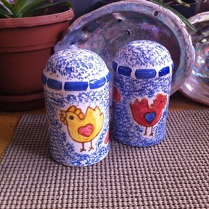 May include: A pair of ceramic salt and pepper shakers. The shakers are cylindrical with blue and white speckled patterns. Each shaker features a cartoon chicken illustration in yellow or red with a heart. A blue band encircles the top.