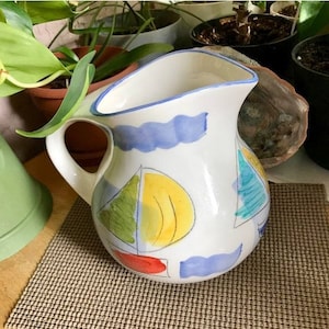 May include: White ceramic pitcher with a curved handle and spout. The pitcher is decorated with blue wave patterns and colorful sailboat designs in yellow, green, and red. The rim is trimmed with blue.