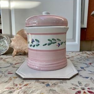 May include: A ceramic canister with a pink lid and base, and a white band with a floral design. The canister sits on a white octagonal tray. The background includes a window and a seashell.