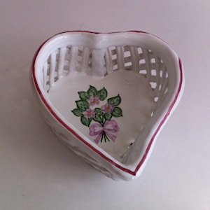 May include: White ceramic heart-shaped basket with a lattice design. The rim is outlined in red. The interior features a floral bouquet with pink flowers, green leaves, and a pink bow. A decorative piece for the home.