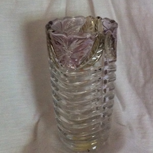 May include: A clear glass vase with a floral design at the top and a wavy, ribbed pattern on the body. The vase has a slight purple tint at the top and is likely used for displaying flowers. The vase is made of glass.