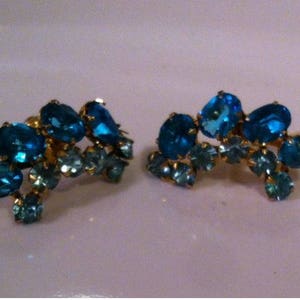 May include: A pair of vintage clip-on earrings featuring clusters of blue and teal-colored rhinestones set in a gold-tone metal. The earrings have an ornate, floral-like design, with various sizes of stones.