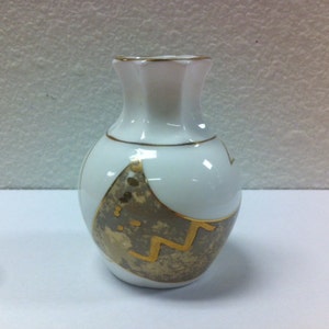 May include: A small, white ceramic vase with a gold rim and decorative gold accents. The vase features a rounded body and a flared, scalloped top. A gold geometric design adorns the front of the vase.