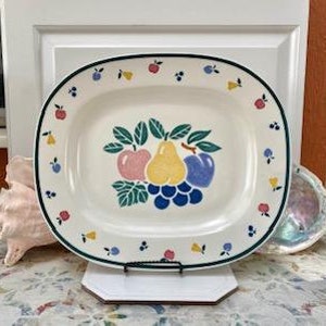 May include: Oval ceramic serving platter with a fruit design. The platter features a central arrangement of a pink apple, yellow pear, and blue plum, with a cluster of blue grapes. The border is decorated with small fruit motifs and a green trim.