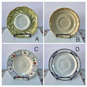 May include: Four decorative saucers are displayed, each with a unique design. Saucer A features a green and white floral pattern. Saucer B is a solid tan color. Saucer C has a blue and red floral design. Saucer D has a blue border.