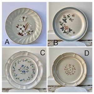 May include: Four vintage ceramic plates with floral designs. Plate A features a beige background with a flower and leaf design. Plate B has a blue rim and floral pattern. Plate C has a scalloped edge and floral design. Plate D has a beige background and floral design.