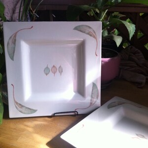 May include: Two white square plates with a leaf design. Each plate has a central recessed area. The plates are decorated with a watercolor-style leaf design in shades of green, brown, and orange.