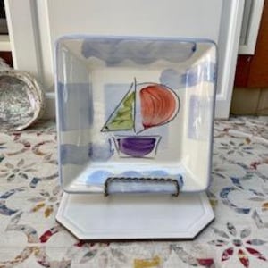 May include: Square ceramic dish with a sailboat design. The dish features a white base with a blue border and a central illustration of a sailboat with green, orange, and purple accents. The dish is displayed on a white stand.