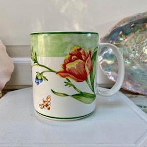 May include: White ceramic mug with a floral design featuring a red and yellow tulip, green leaves, and a small bee. The mug has a green rim and handle, and is set against a blurred background.