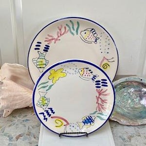 May include: Two white ceramic plates with a blue rim, featuring hand-painted fish and coral designs in various colors. The plates are of different sizes, with the larger one measuring approximately 10 inches in diameter. The plates are part of a nautical or coastal-themed dinnerware set.