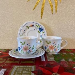 May include: Two white teacups and saucers with a floral design featuring pink, blue, and yellow flowers. The teacups have handles and sit on matching saucers. A third saucer is behind the cups. The background is a textured cream color.