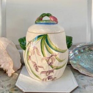 May include: A cream-colored ceramic jar with a lid and handles, adorned with a floral design in shades of purple, green, and blue. The jar has a woven handle and is set on a white surface.